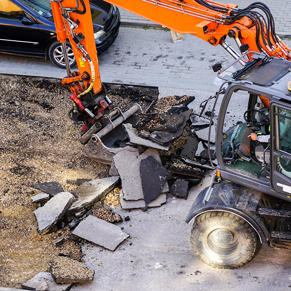 An excavator breaks up the old asphalt layer with a bucket during street repairs in city